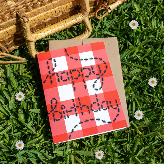 Birthday card with a red and white checker print and graphic that reads "happy birthday" spelled out with drawing of ants, on a grass background.