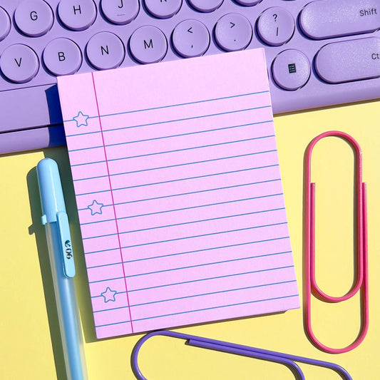 pink notepad with lined pages, blue pen, pink paper clip on a yellow background with purple keyboard.