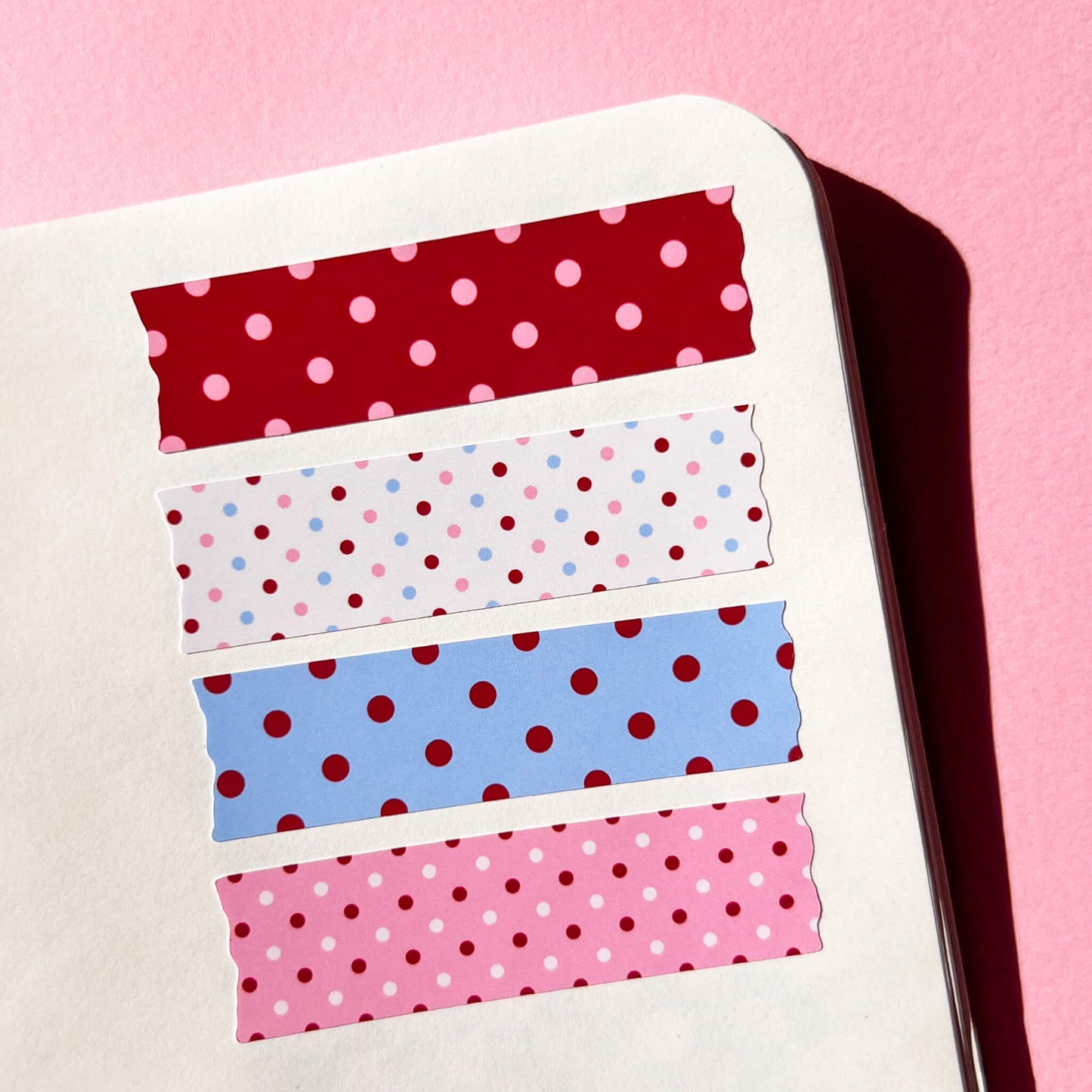 Four strips of decorative washi tape stickers with polka dot patterns arranged vertically on a white notebook page against a solid pink background.