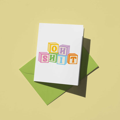 Card with colorful block letters spelling 'OH SHIT' on a green envelope against a beige background