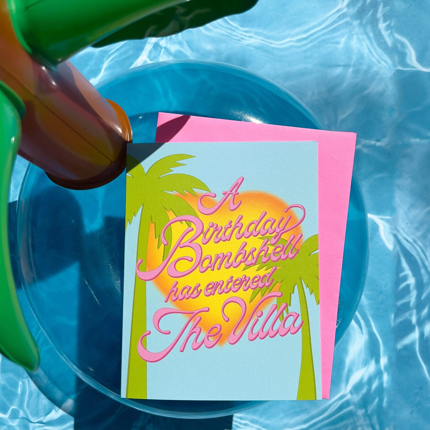 Birthday card with palm trees and 'A Birthday Bombshell has entered The Villa' text, pink envelope on a inflatable pol floatie.