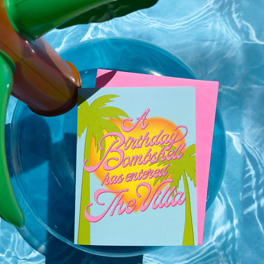 Birthday card with palm trees and 'A Birthday Bombshell has entered The Villa' text, pink envelope on a inflatable pol floatie.