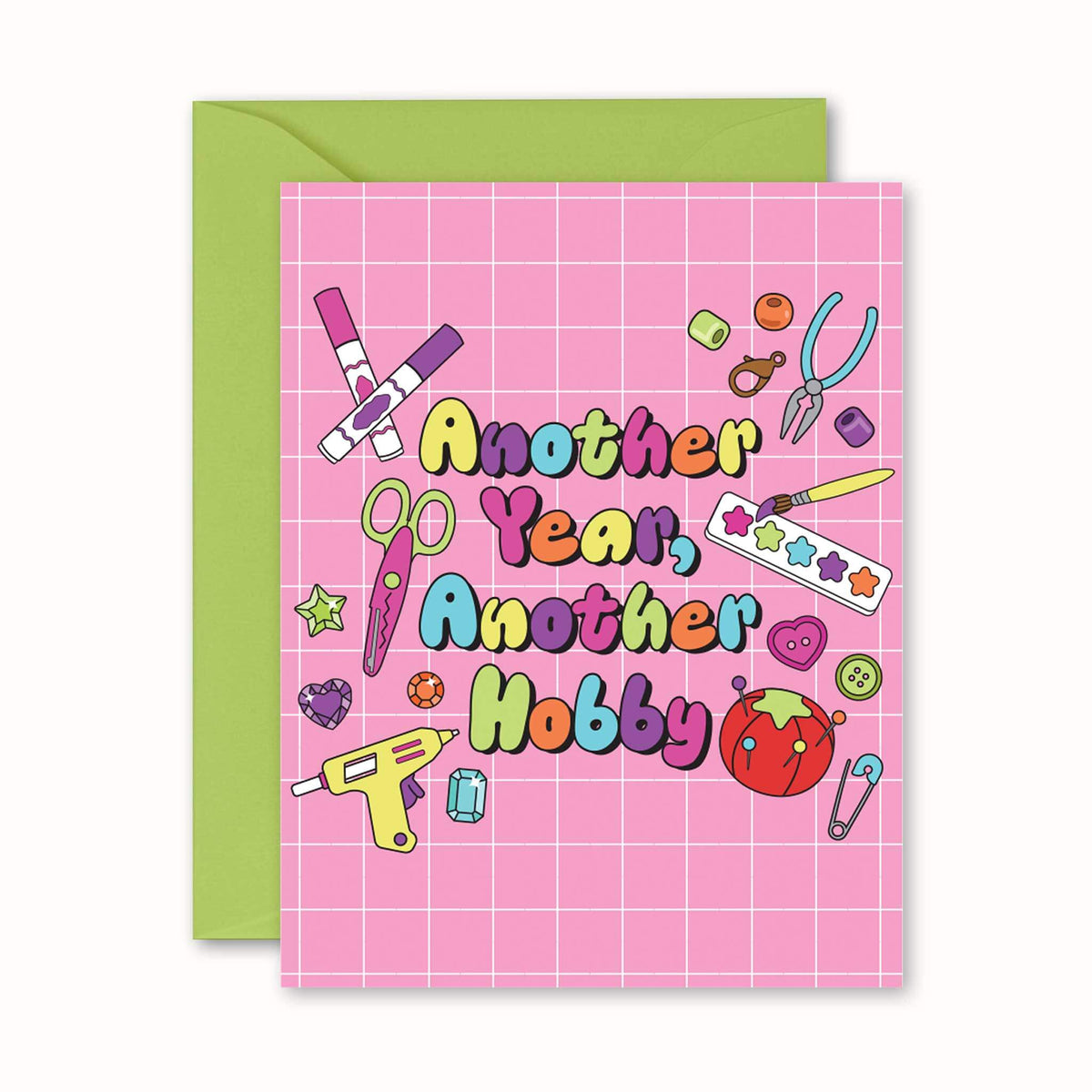 A pink greeting card with a grid pattern and colorful text reading 'Another Year, Another Hobby,' shown with a green envelope behind it on a white background.