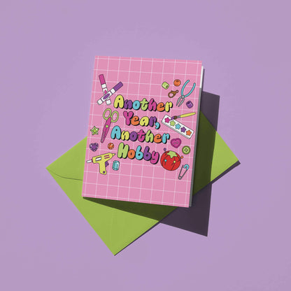 A pink grid-patterned greeting card with the text 'Another Year, Another Hobby' in colorful bubble letters, styled on top of a green envelope against a solid lavender background.