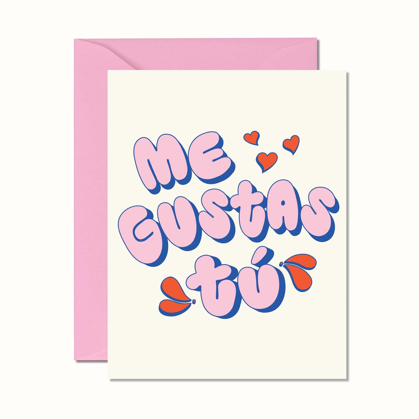 A greeting card with a pink envelope behind it, featuring the Spanish phrase 'Me GUSTAS tú' in bubbly pink letters with blue outlines and small red hearts on a cream cardstock.