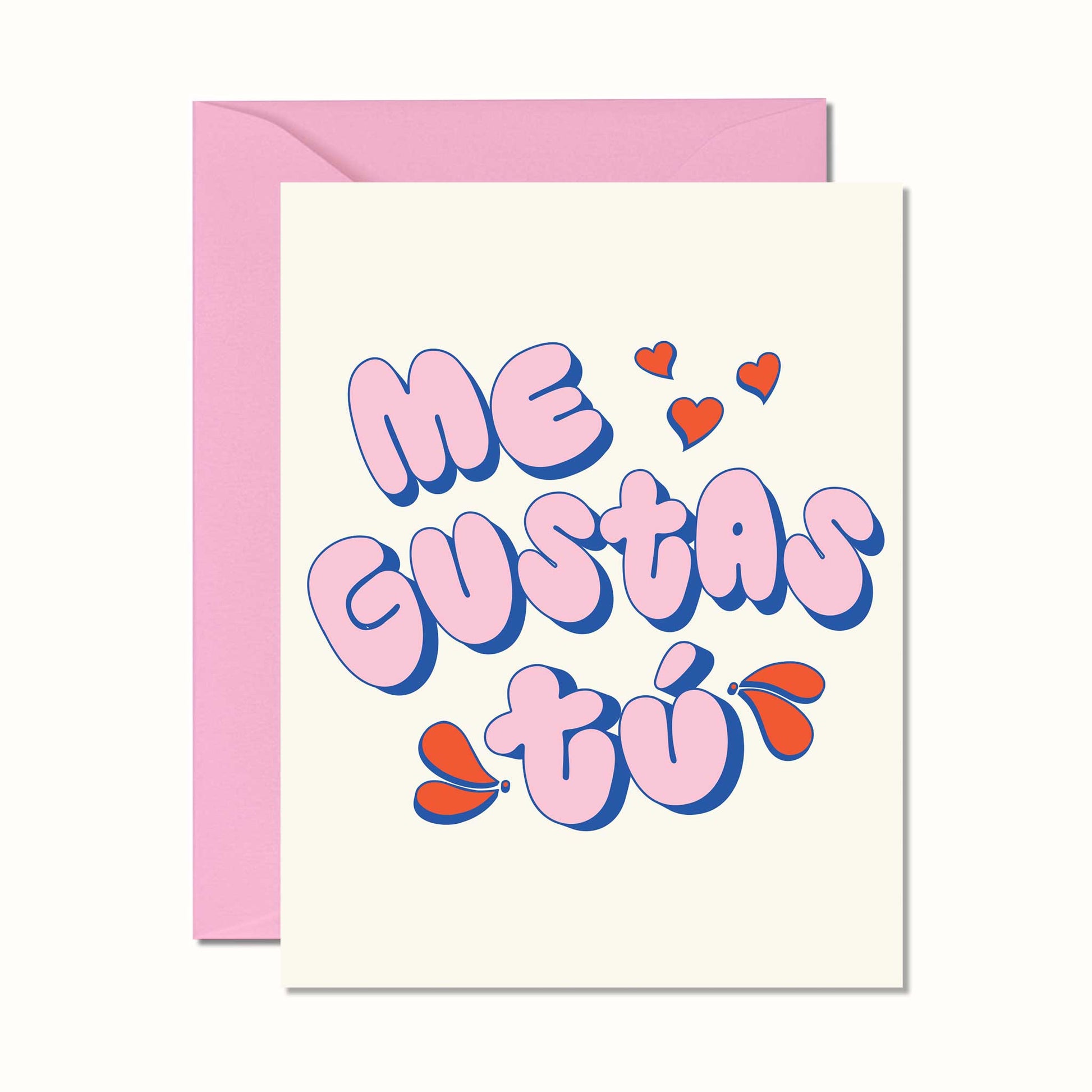 A greeting card with a pink envelope behind it, featuring the Spanish phrase 'Me GUSTAS tú' in bubbly pink letters with blue outlines and small red hearts on a cream cardstock.