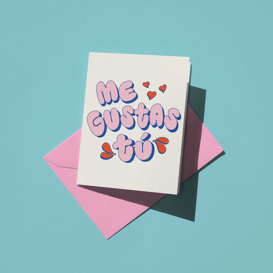 A white greeting card with the Spanish phrase 'ME GUSTAS TÚ' in bubbly pink and blue lettering, accented with small red hearts, resting on top of a pink envelope against a solid light blue background.