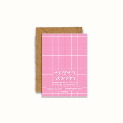 The back side of a pink card with a white grid pattern and the text 'No Year For Sure', shown with a brown kraft envelope behind it on a light cream background.
