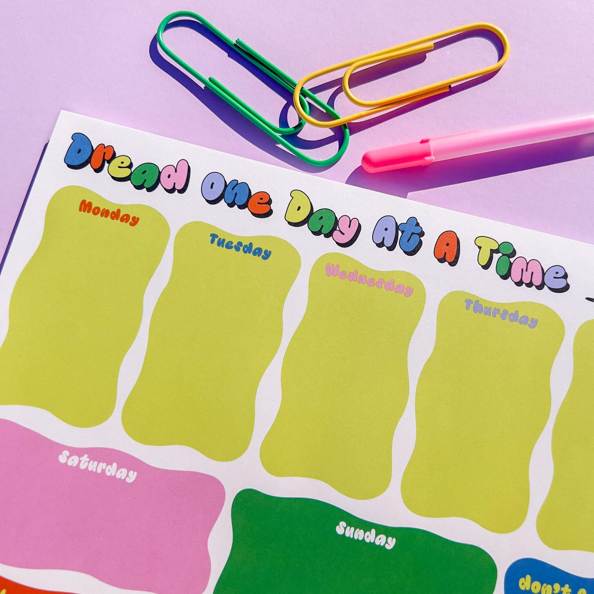 Weekly planner with days of the week on a purple background with colorful paper clips and a pink pen.