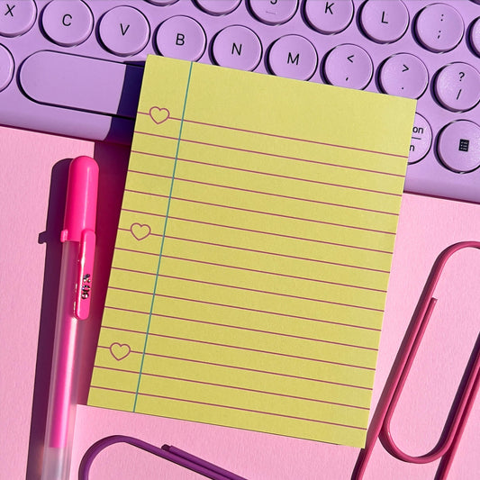 Yellow notepad with pink pen and paper clips on a pink surface with purple keyboard