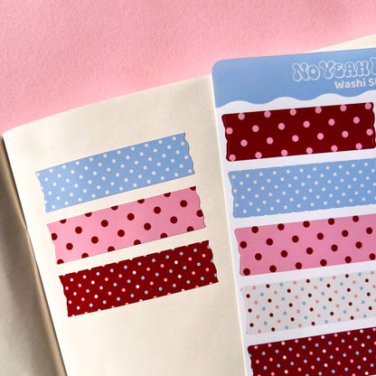 A close up of a washi tape sticker sheet showing several patterned strips, including blue and pink polka dots displayed next to an open notebook on a pink background.