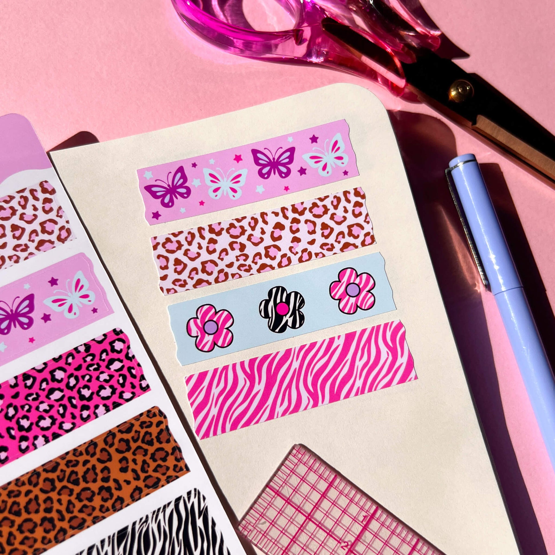 A styled flat-lay of decorative washi tape sticker sheets with pink leopard, butterfly, flower, and pink zebra patterns, arranged on a pink surface with scissors, a pen, and a ruler nearby.