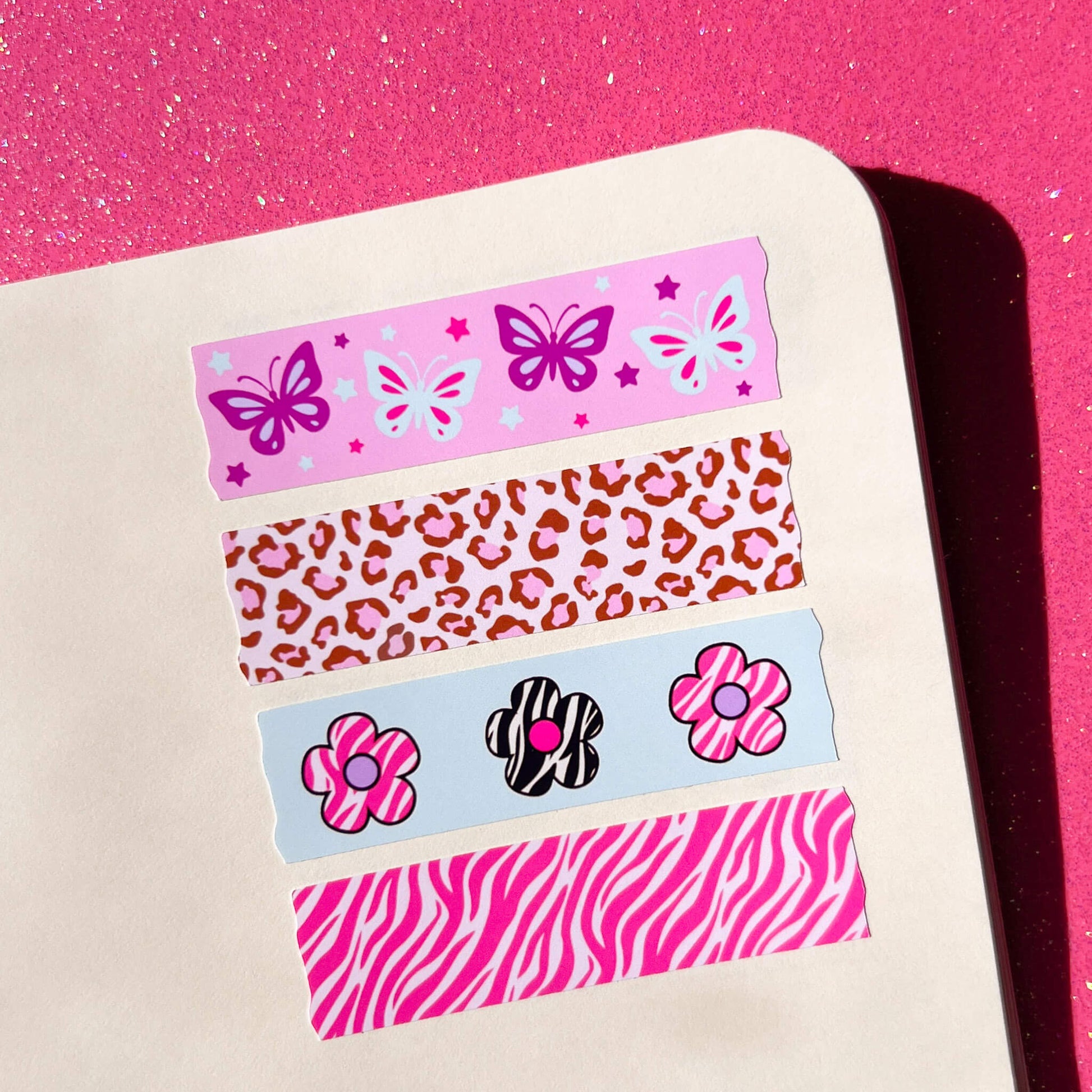 Four decorative washi tape stickers with pink patterns (butterflies, leopard print, flowers, zebra stripes) arranged on a cream colored notebook against a bright pink glitter background.