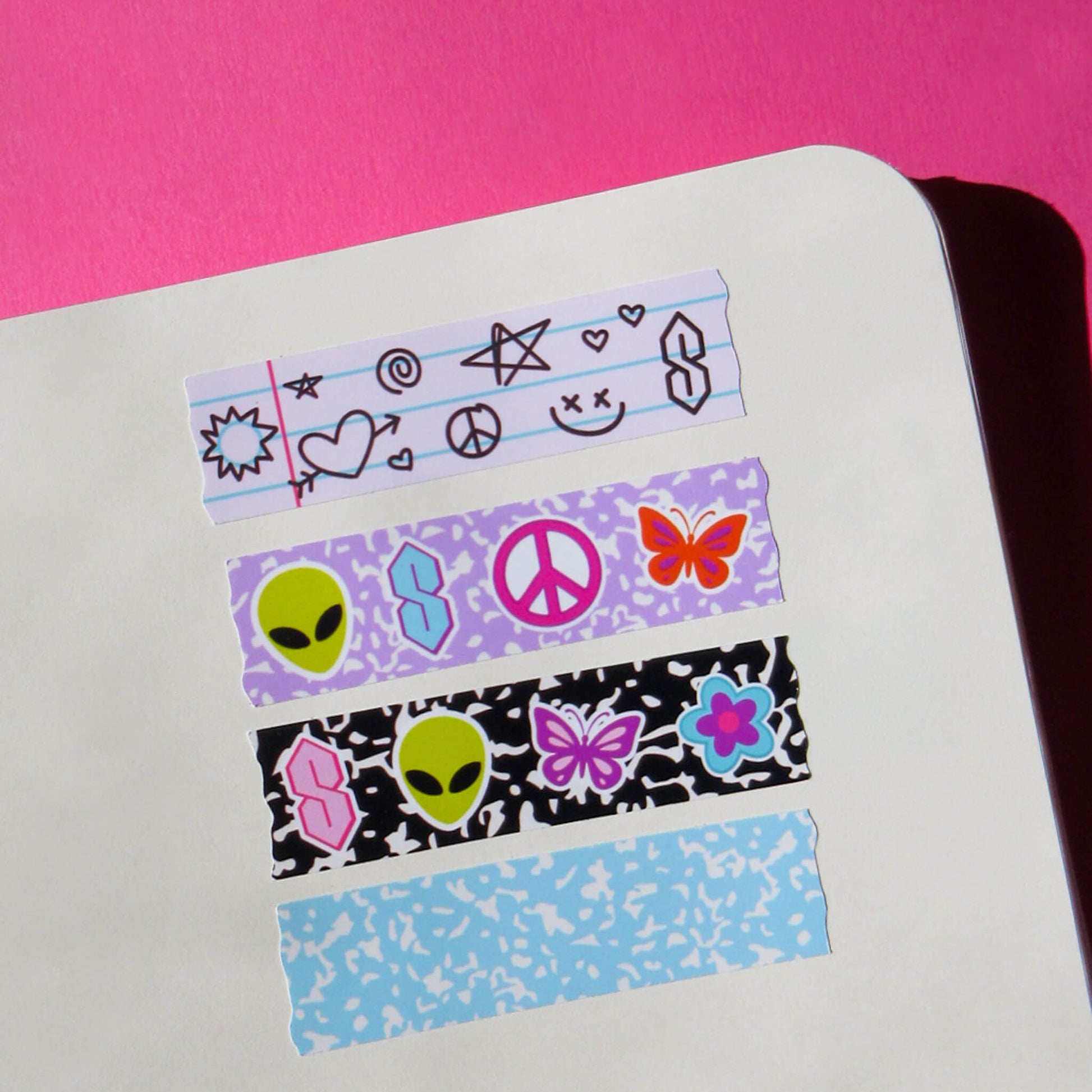 Four strips of decorative washi tape stickers with retro y2k themed designs, including aliens, peace signs, butterflies, and stars, arranged on a white notebook page against a bright pink background.