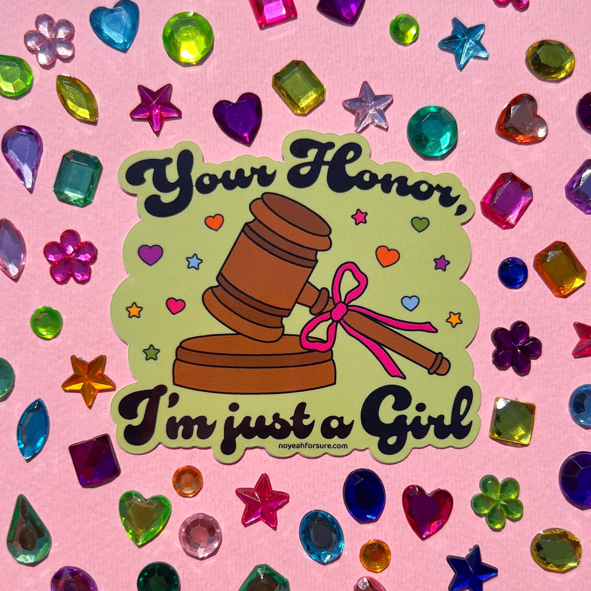 Sticker featuring a judge's gavel with a pink bow and the whimsical text, "your honor, i'm just a girl" surrounded by colorful hearts and stars on a pink background with gems. 